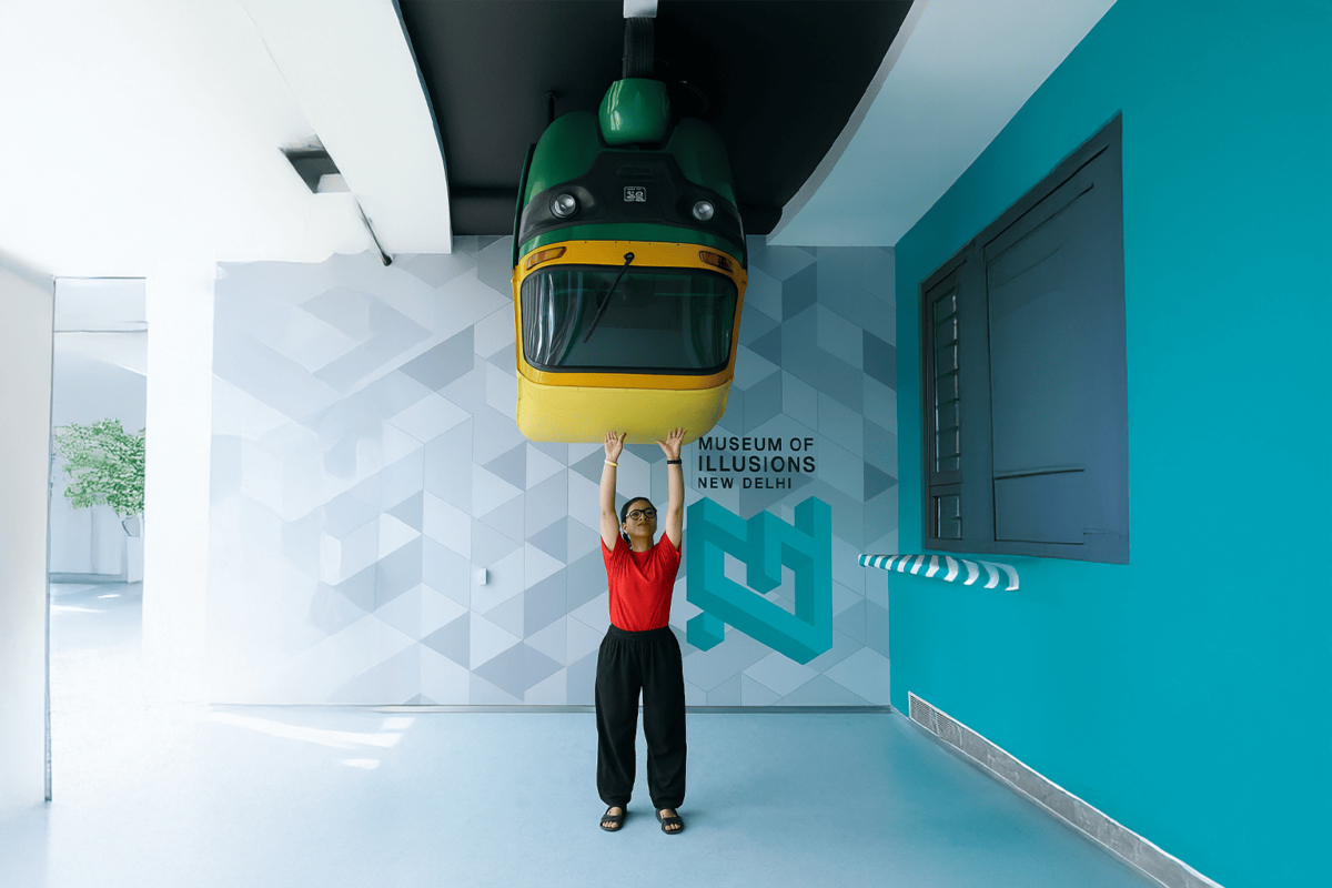Image of a young Indian woman in a red t-shirt appearing to support an upside-down auto-rickshaw at the Museum of Illusions, New Delhi. The illusion takes place against a teal geometric-patterned wall with the museum’s logo, perfectly aligning with the brand’s signature color palette.